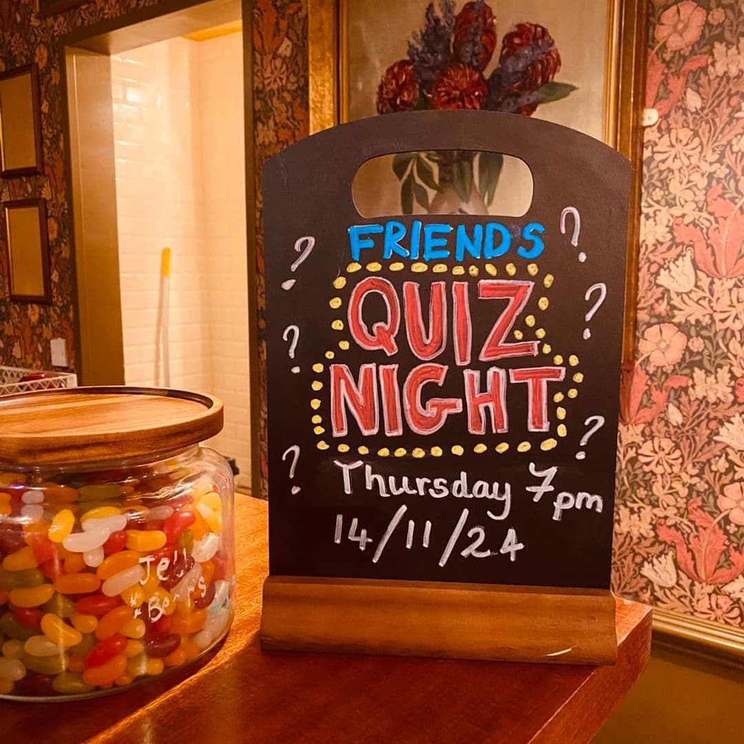 Chalkboard announcing 'Friends Quiz Night' event at Hesco Lounge on Thursday at 7 pm, with a jar of jelly beans in the foreground and decorative wallpaper in the background.