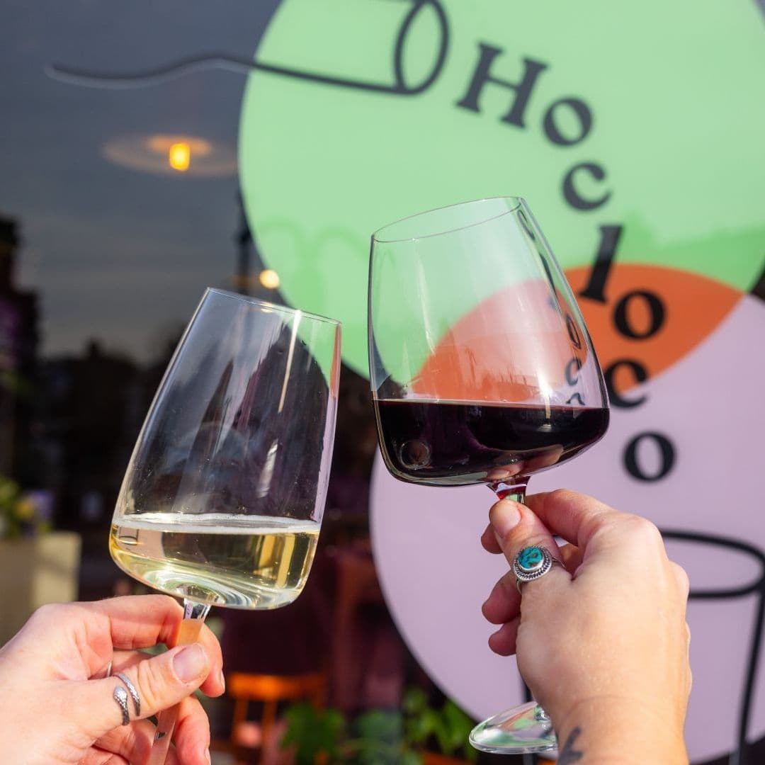 Two hands toasting with wine glasses in front of the Hoc Loco sign, one glass with white wine and the other with red wine.