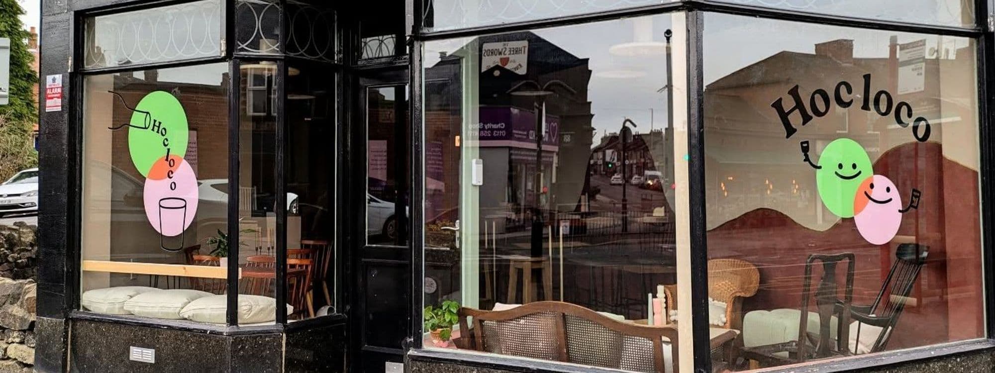Exterior view of Hoc Loco with large windows displaying colorful graphics of smiling characters, one holding a wine glass and the other a phone, reflecting the street scene.