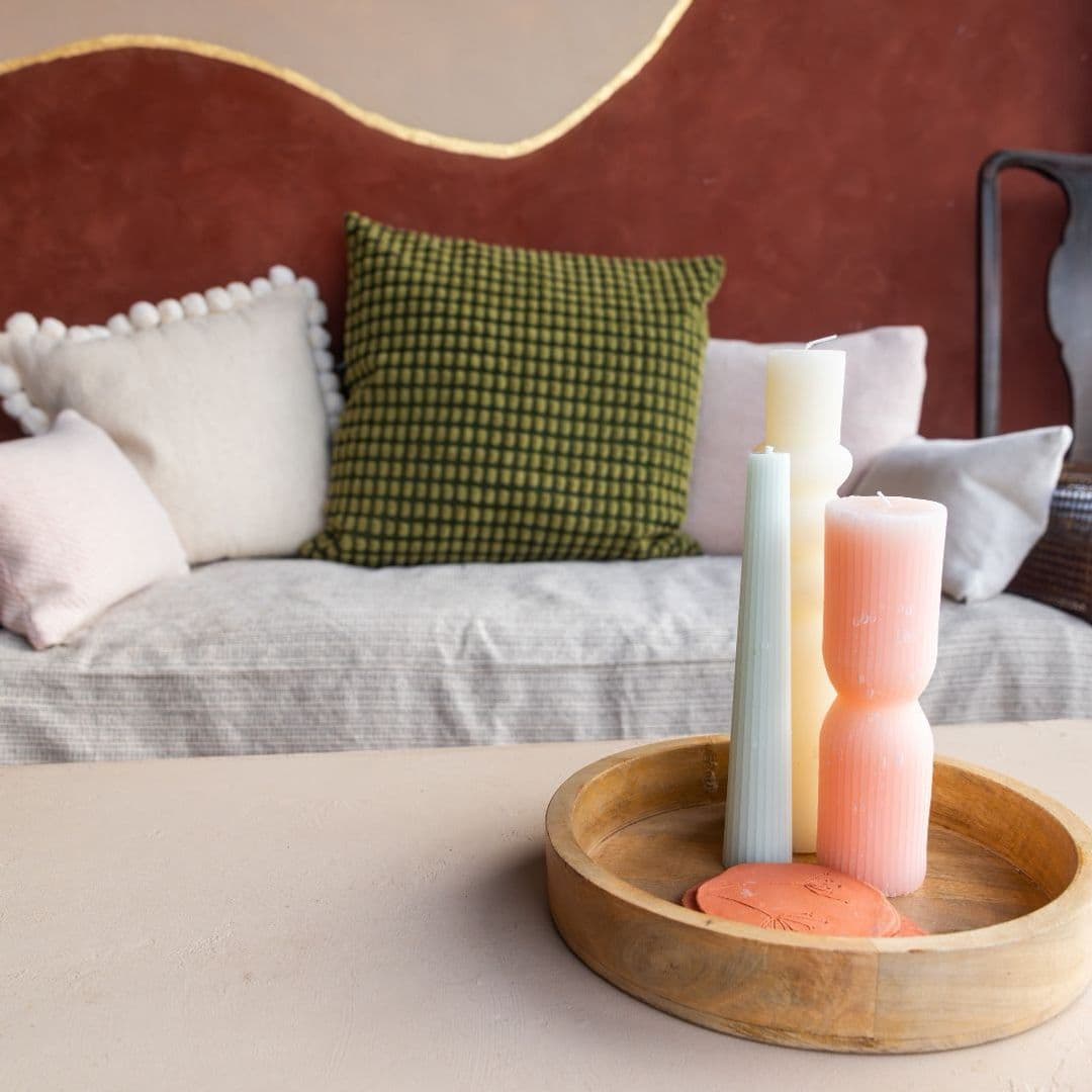 Cozy corner at Hoc Loco with a rustic wooden tray holding pastel-colored candles, set against a backdrop of plush pillows on a textured bedspread and a terracotta-colored wall with decorative molding.