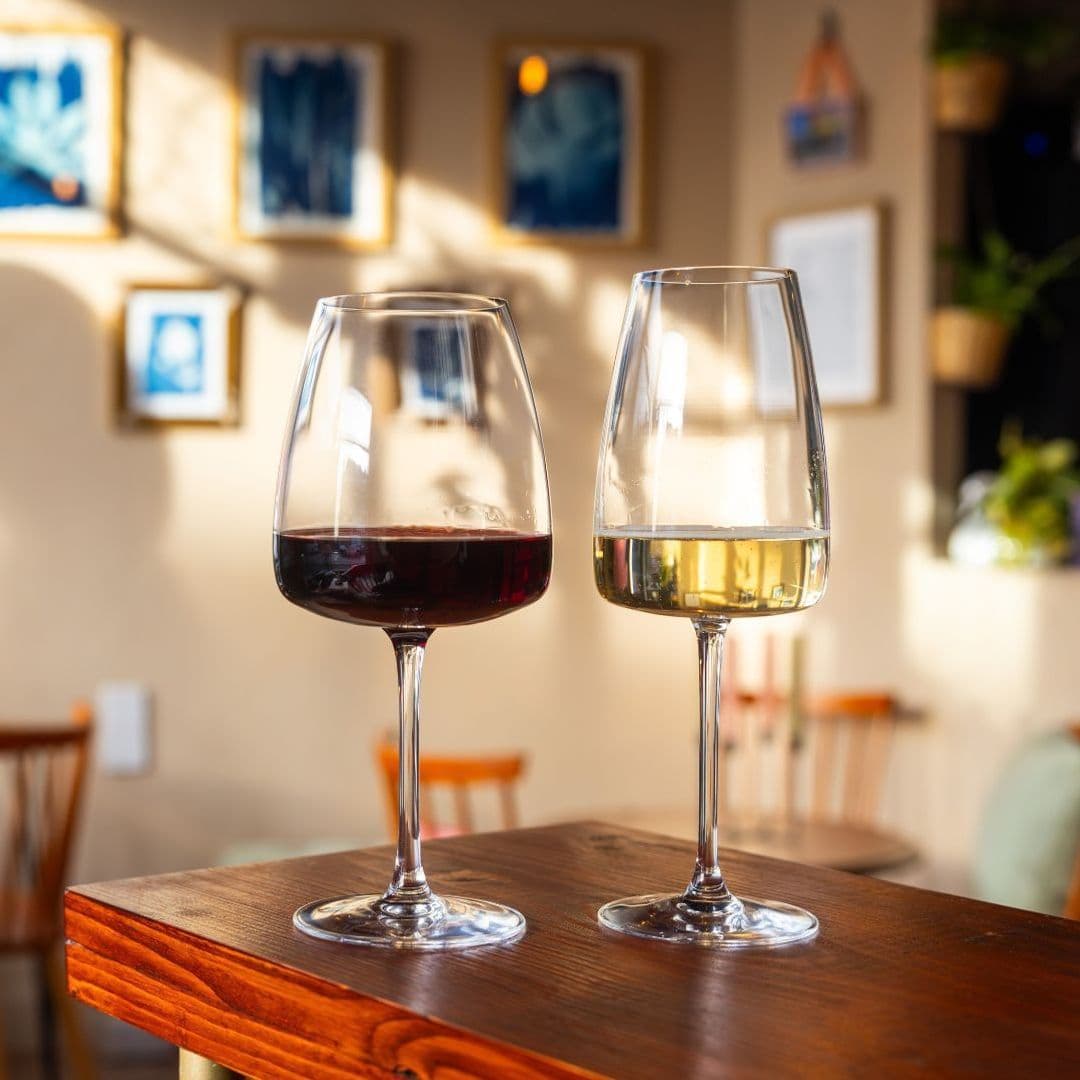 Two glasses of wine on a wooden table at Hoc Loco, with a glass of red wine on the left and white wine on the right, in a cozy dining setting with warm lighting and art on the walls.