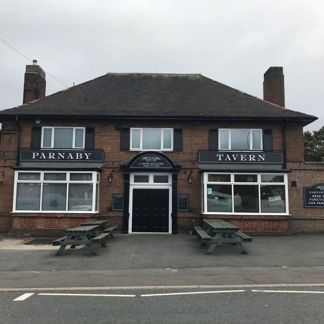 Exterior view of Parnaby Tavern, a traditional brick building with a black and white signboard, featuring a central entrance flanked by windows labeled 'Bar' and 'Lounge', and outdoor benches in front.