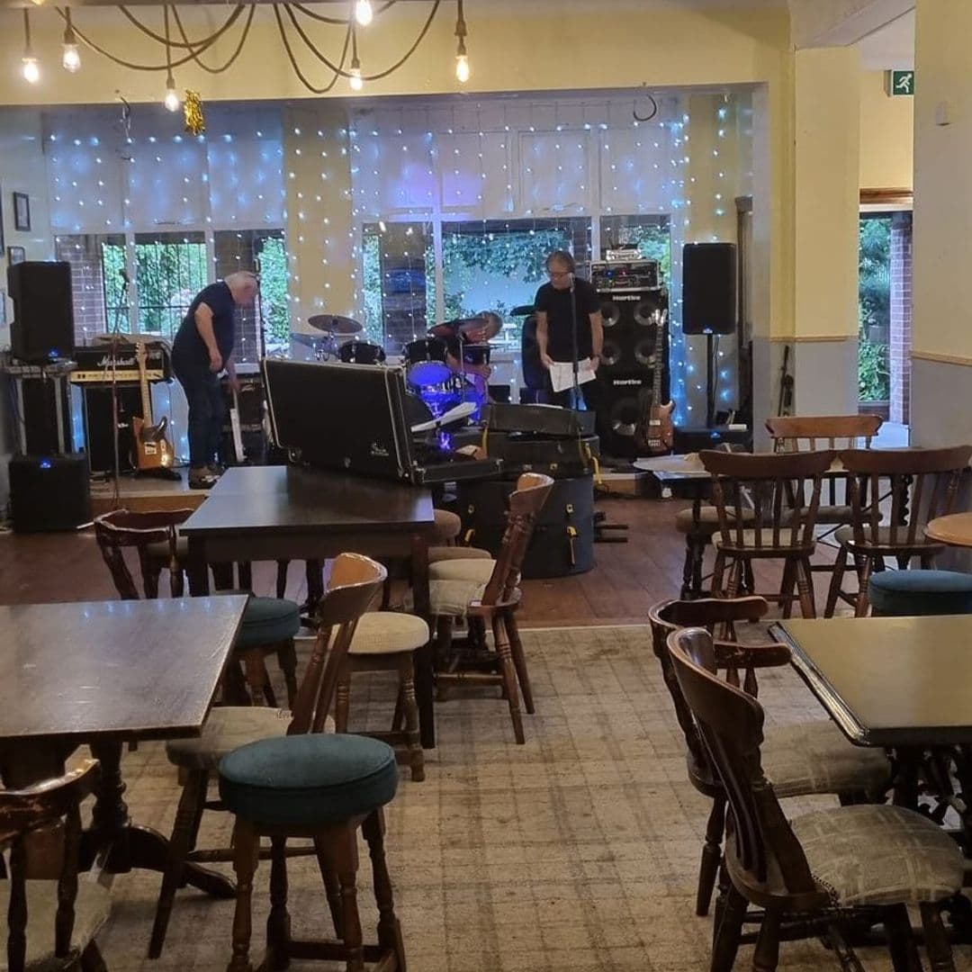 Interior of Parnaby Tavern with a live music setup including speakers, a drum set, and guitars, with string lights above the window and wooden tables and chairs in the foreground.