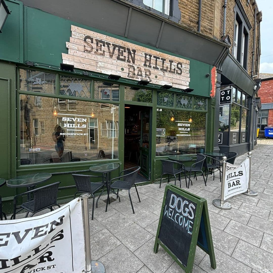 Exterior view of Seven Hills Bar with outdoor seating, a "Dogs Welcome" sign, and visible interior through the open doorway.