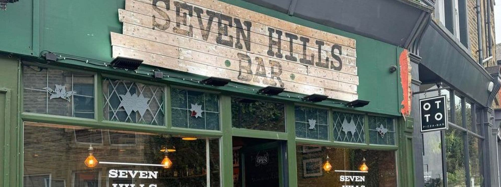 Exterior view of Seven Hills Bar with rustic wooden signage and decorative window stars, adjacent to OTTO Pizza Bar.