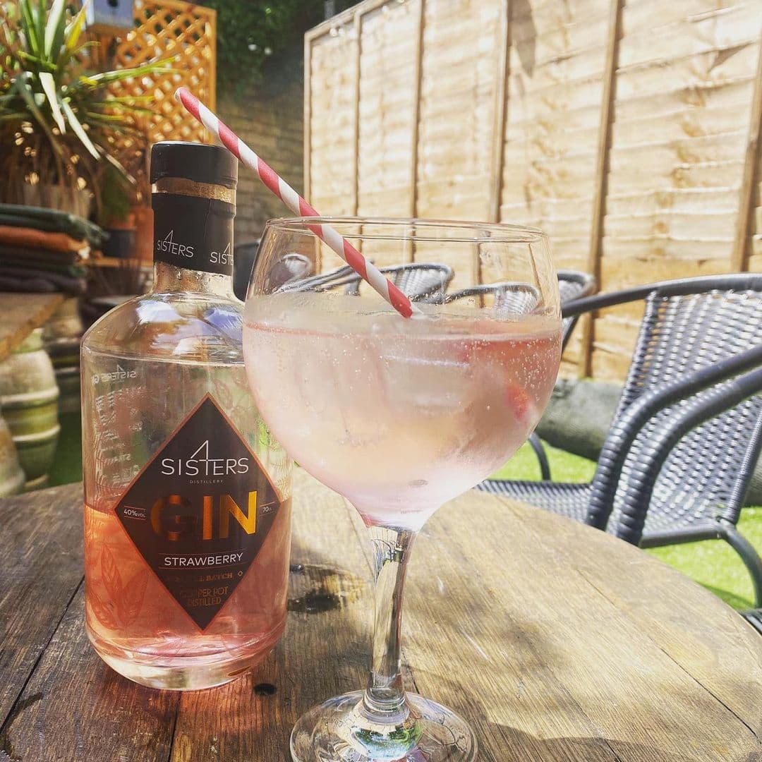 Bottle of strawberry gin and a gin cocktail with ice and a striped straw on a wooden table at Seven Hills, with garden chairs and a wooden fence in the background on a sunny day.