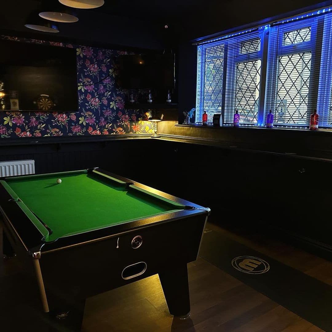 Interior of The Commercial featuring a pool table with a green felt surface, under ambient lighting, with colorful floral wallpaper and stained glass windows in the background.