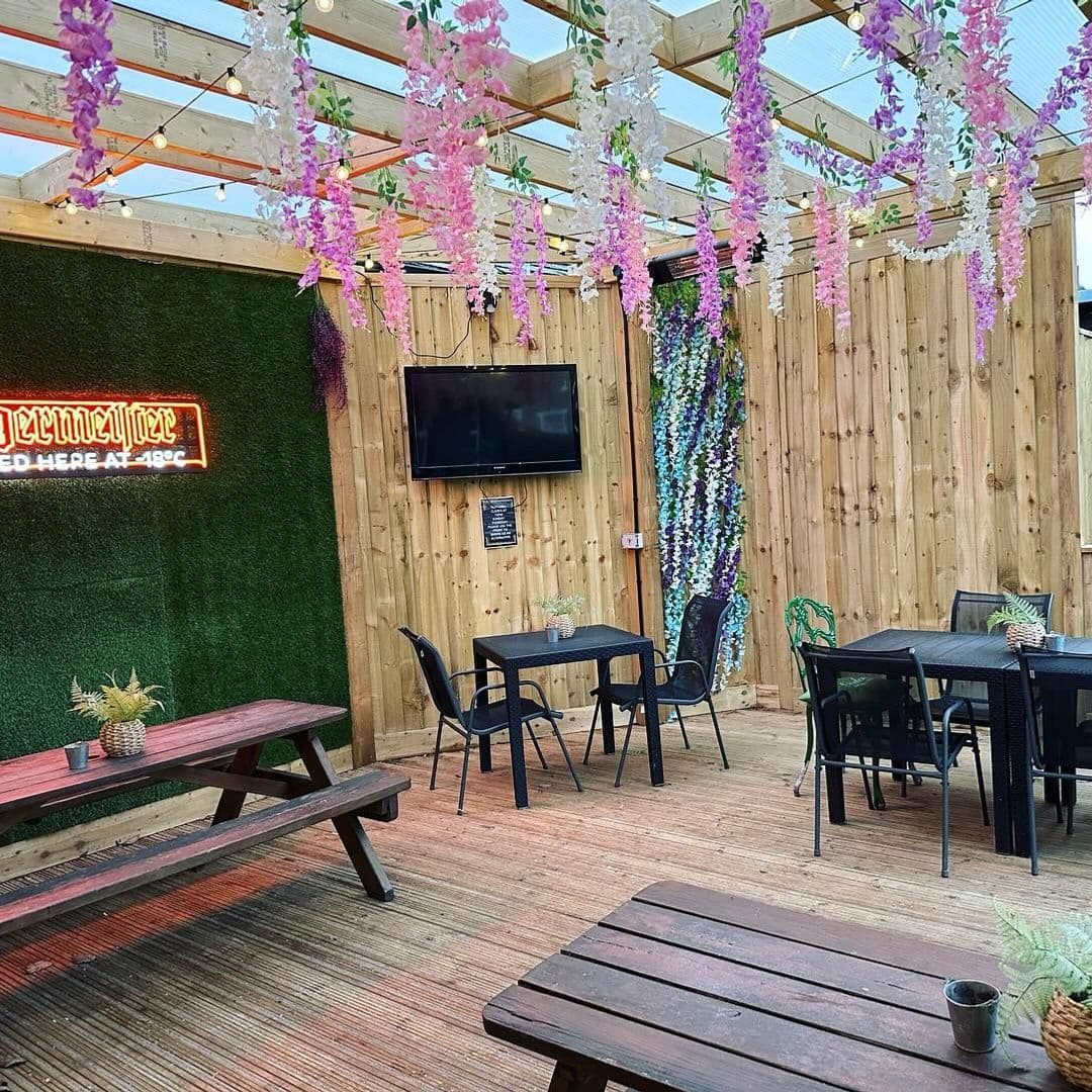 Cozy outdoor seating area at The Commercial with wooden benches, tables, and chairs, adorned with hanging pink and purple flowers, a neon sign, and a wall-mounted TV.