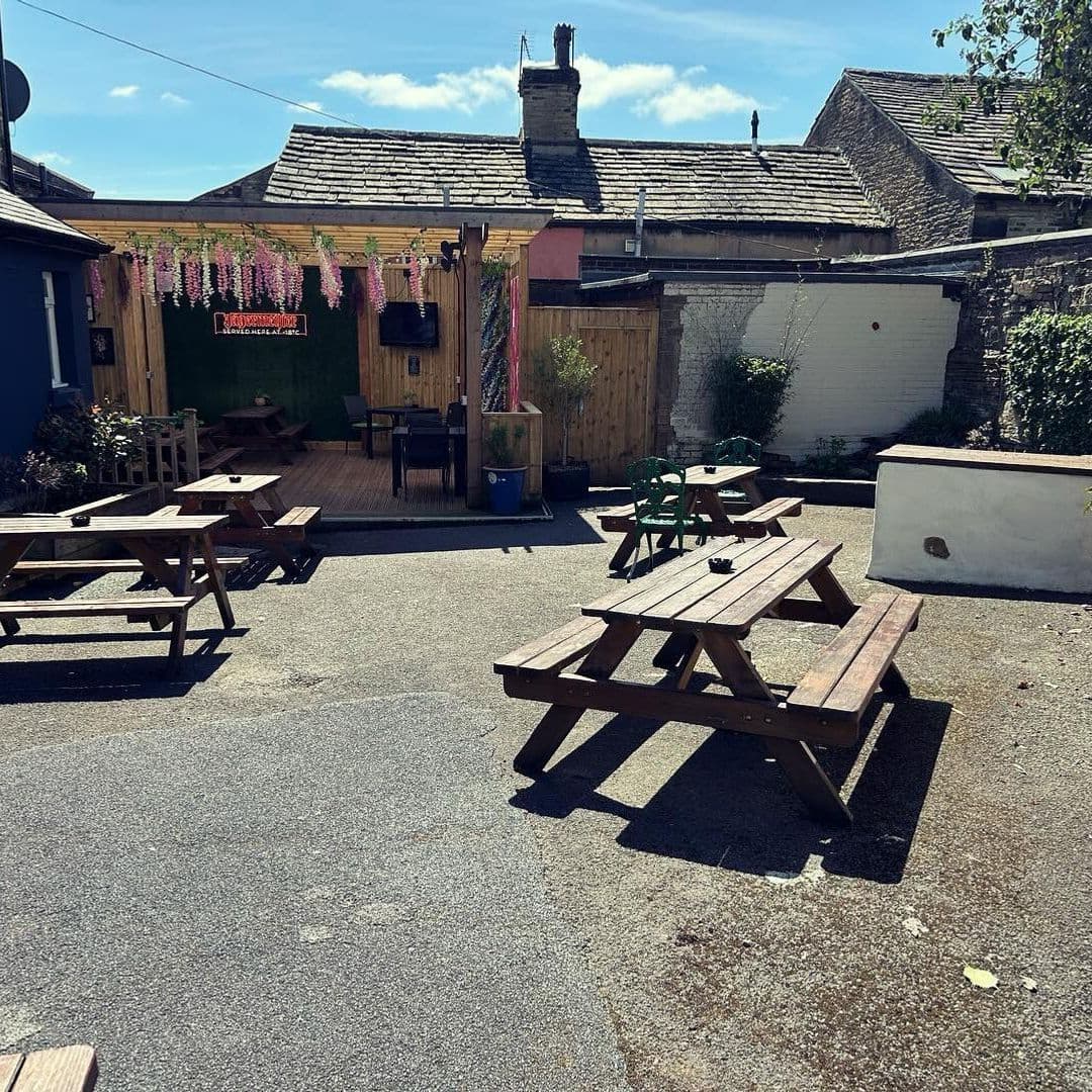 Outdoor seating area at The Commercial with wooden picnic benches, decorative hanging flowers, and a clear blue sky.