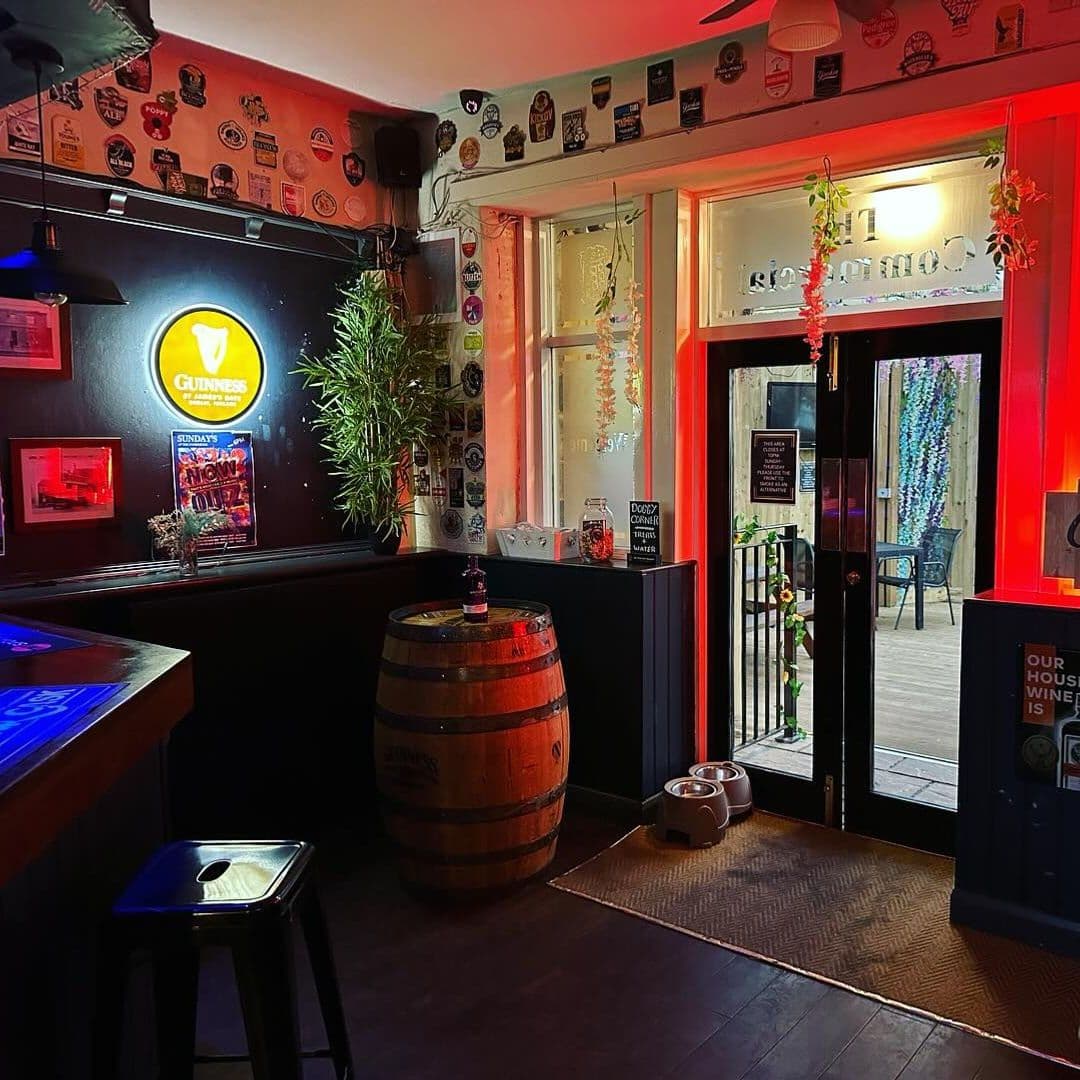 Interior of The Commercial showing a cozy pub atmosphere with warm lighting, a Guinness sign on the wall, a wooden barrel table, and decorative beer coasters above the bar.