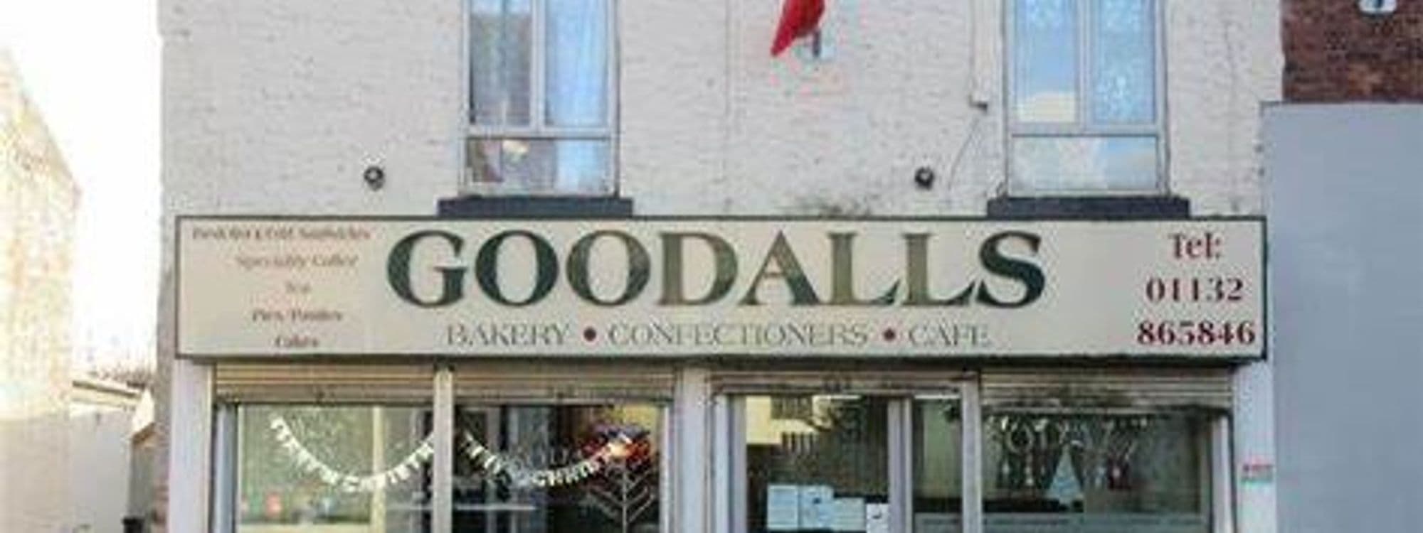 Alt text: Exterior view of Goodalls Bakery, featuring a sign with the name "Goodalls" and offerings like bakery items, confectionery, and café services. A flag is visible above the entrance.