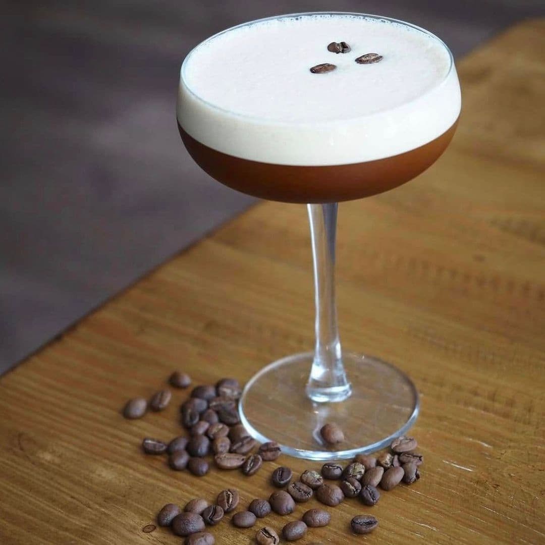 A close-up of a cocktail glass filled with a creamy drink, topped with coffee beans, resting on a wooden table with scattered coffee beans around it. Kofi&Co – Wetherby.