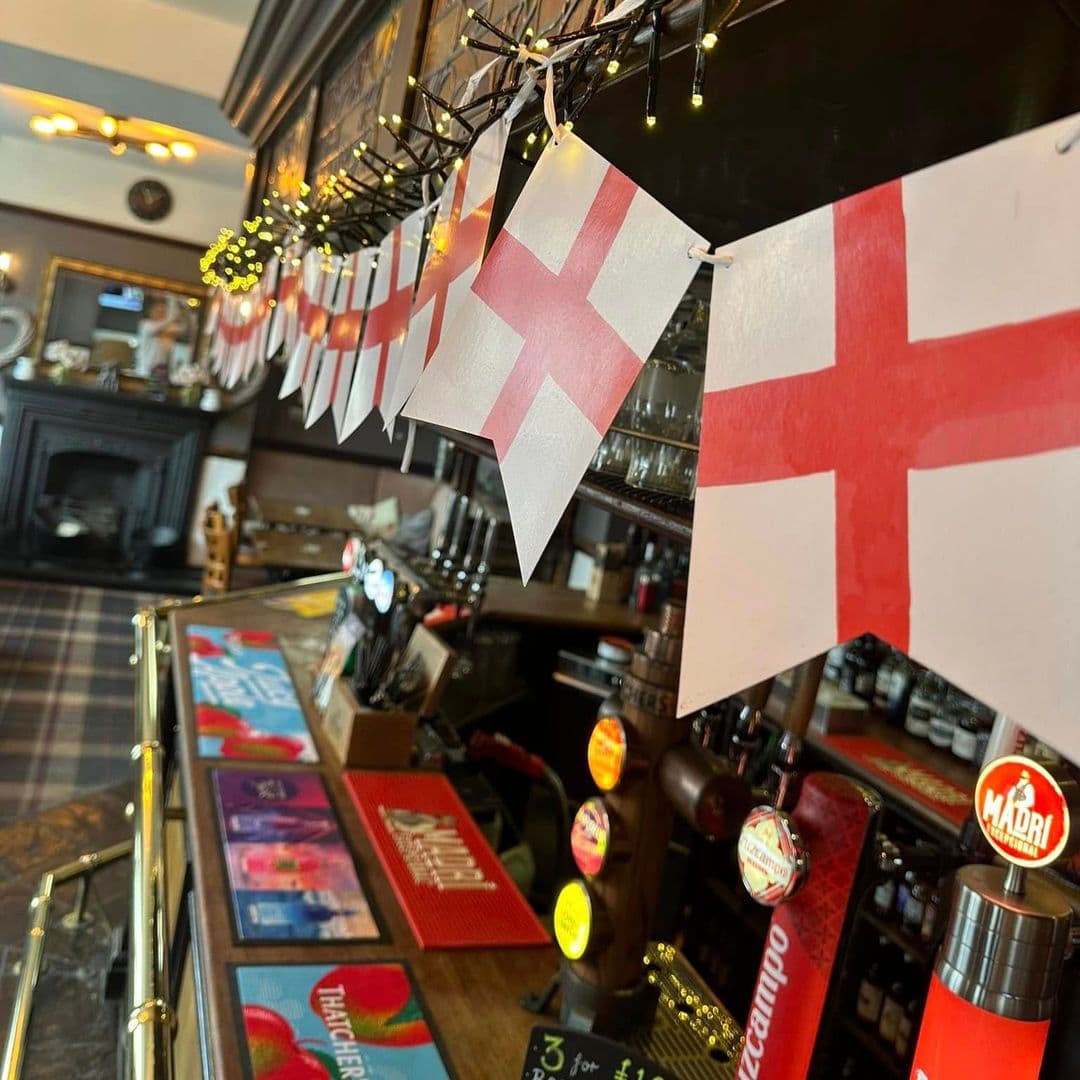 Alt text: Interior view of The Hare and Hounds – Rothwell, featuring a bar decorated with English flags and colorful drink mats. The warm atmosphere includes a fireplace in the background and a variety of beer taps on display.