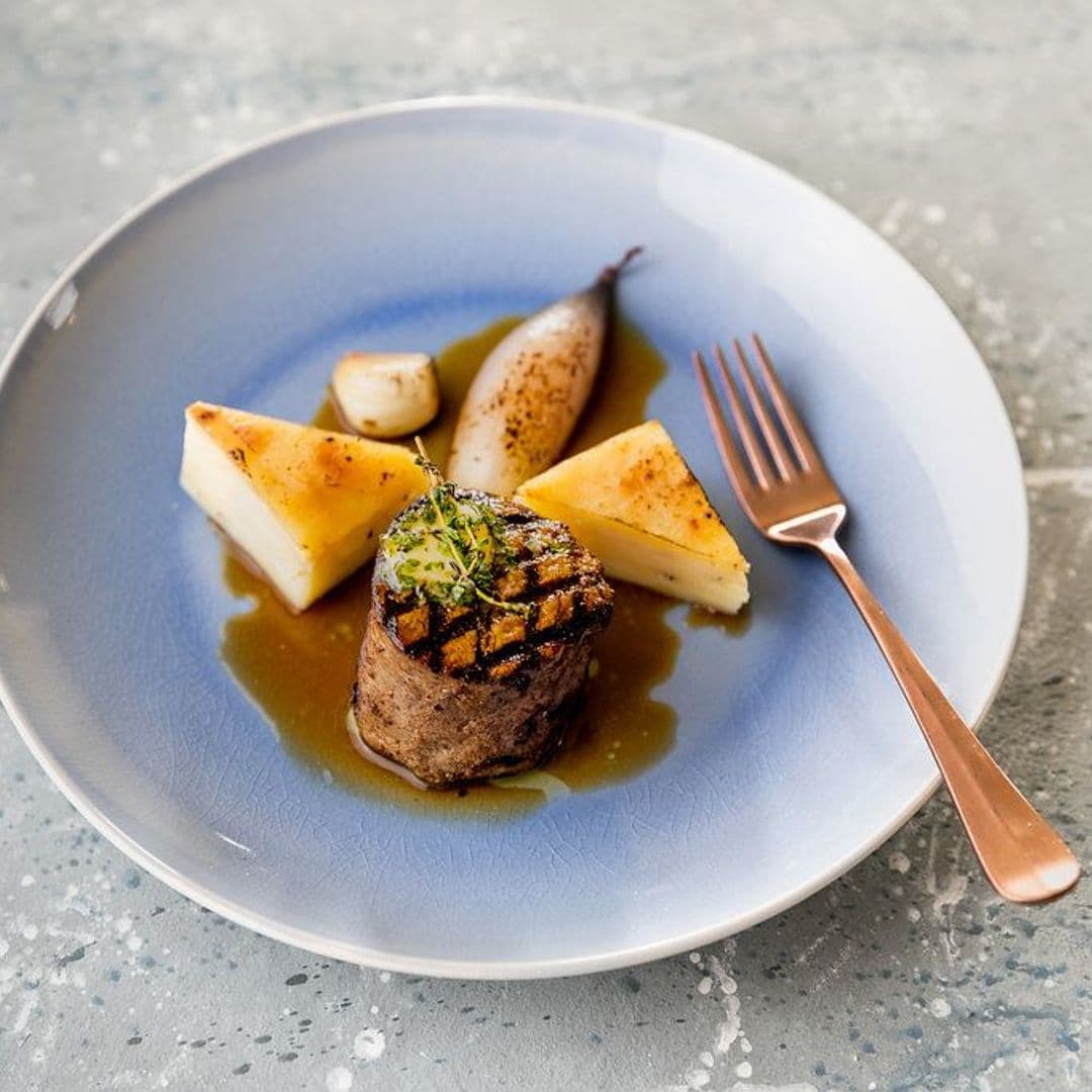 Delicious gourmet dish served at The Mansion, featuring a perfectly grilled piece of meat, accompanied by triangular potato wedges and sautéed vegetables, all presented on a stylish blue plate with a drizzle of sauce. A fork is placed beside the plate, enhancing the elegant dining experience.