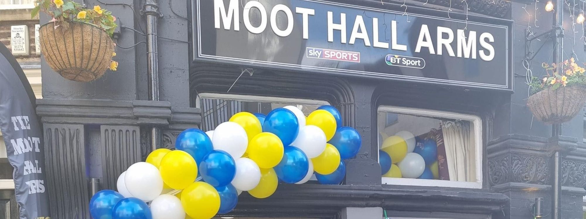 Alt text: The Moot Hall Arms pub entrance decorated with blue, yellow, and white balloons, featuring a sign that reads "Moot Hall Arms." Flower baskets hang from the building, adding a welcoming touch.