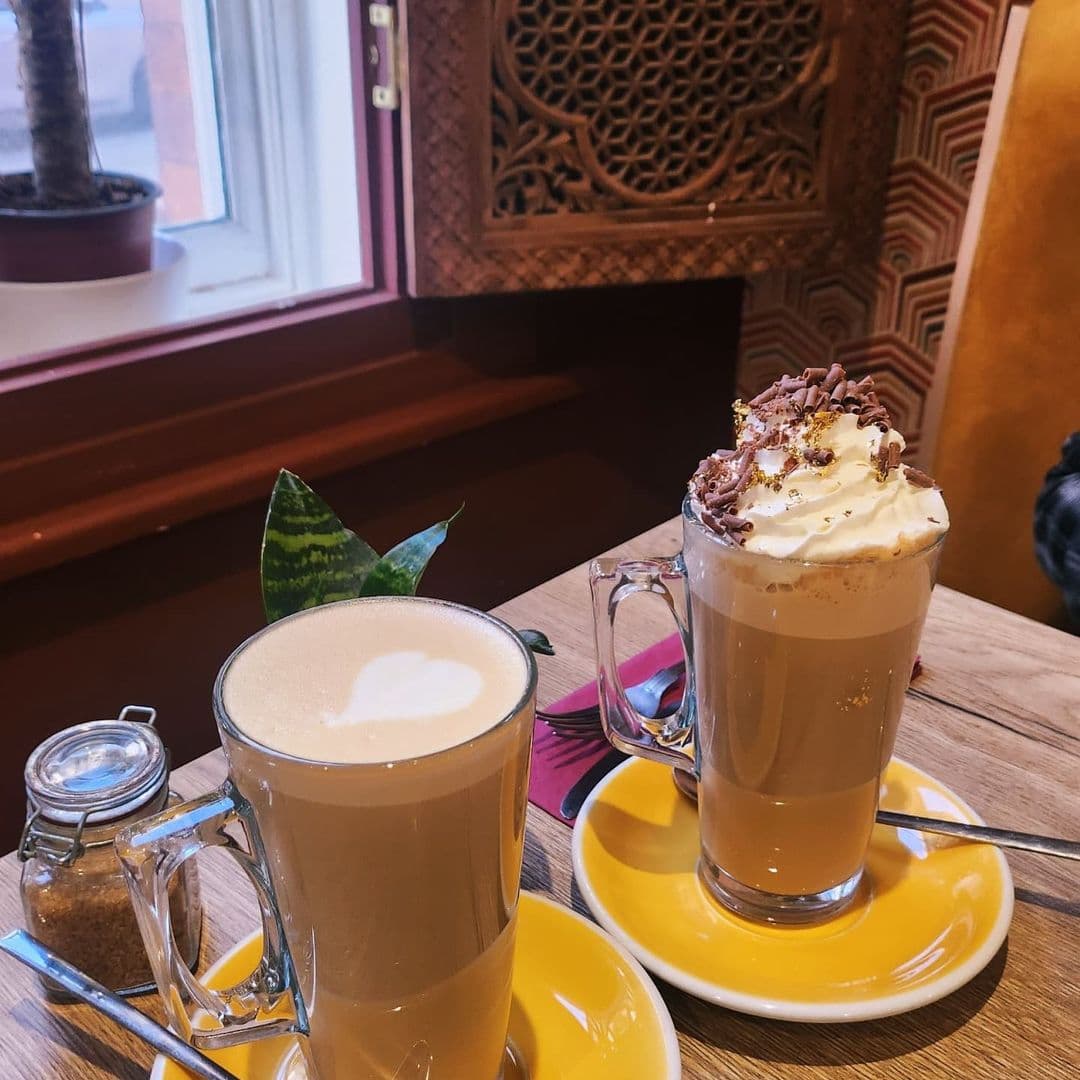 Two delicious coffee drinks are displayed on a wooden table at Wolfox Coffee. One drink features a heart-shaped foam design, while the other is topped with whipped cream and chocolate shavings. A small jar of sugar and a green plant are also visible in the scene.