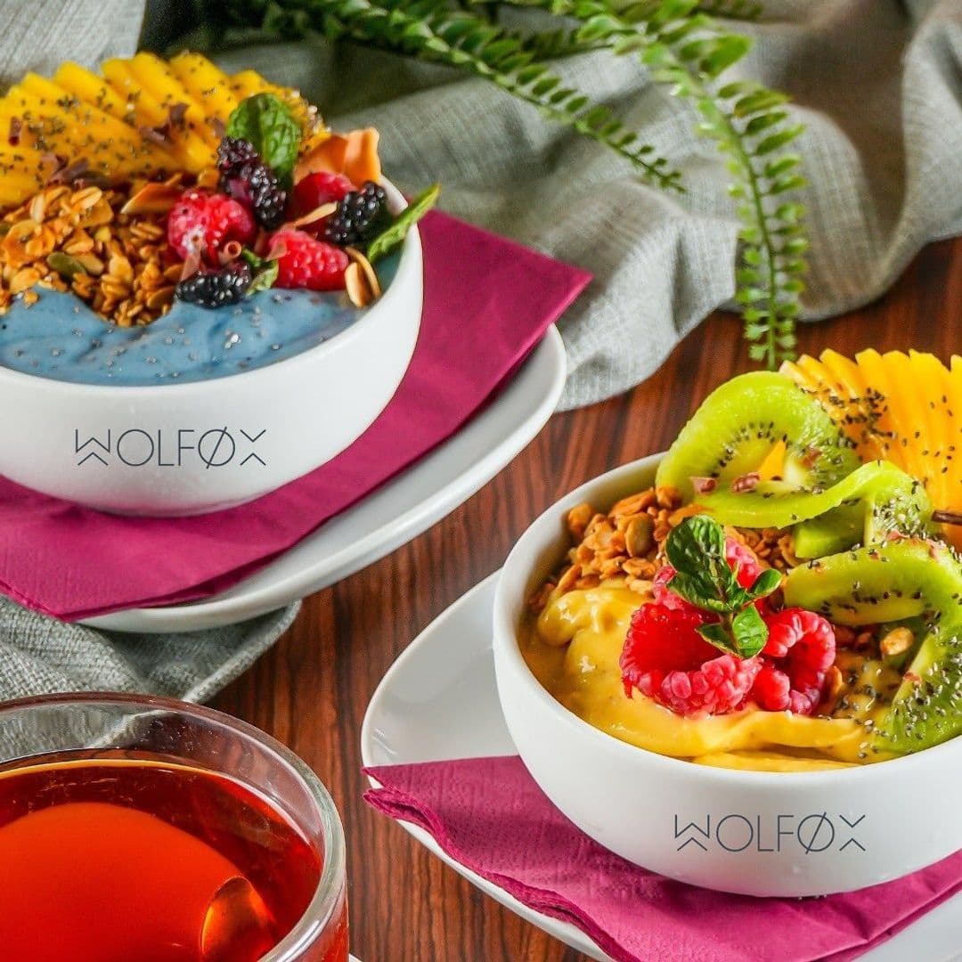 Two colorful smoothie bowls are served at Wolfox Coffee. One bowl features a blue smoothie topped with granola, fresh raspberries, blackberries, and mango slices. The other bowl contains a yellow smoothie topped with kiwi, granola, raspberries, and mint leaves. A cup of tea is placed beside the bowls on a wooden table, with a soft gray cloth in the background.