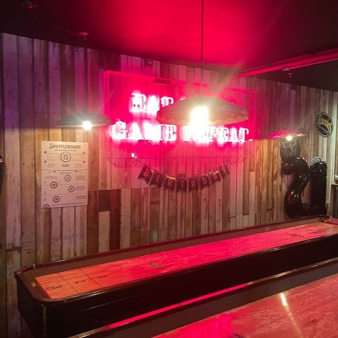 Alt text: Interior of The Head of Steam Headingley featuring a shuffleboard table, warm lighting, and a neon sign that reads "Game Repeat." A birthday banner and decorative balloons add to the festive atmosphere.