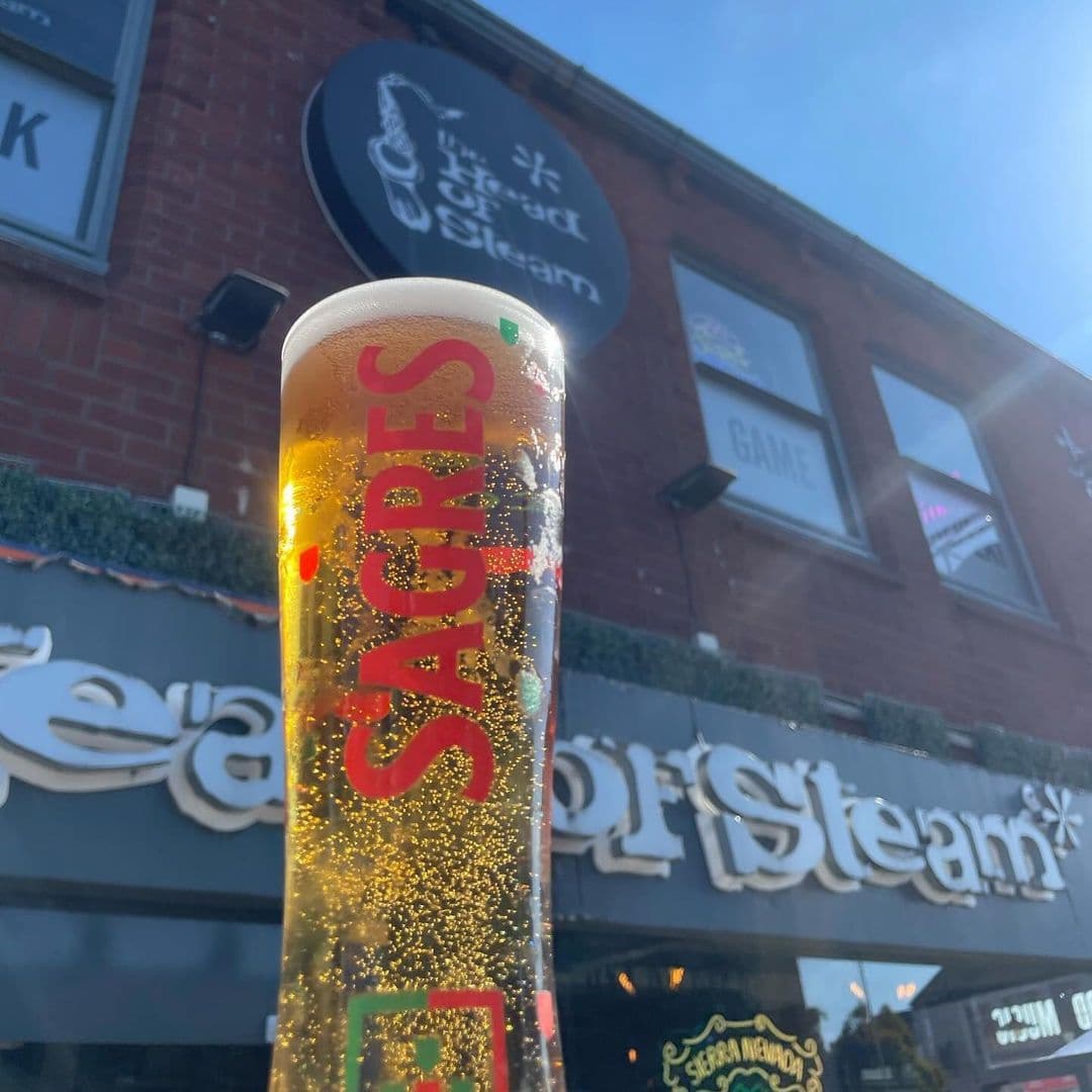 A tall glass of sparkling beer with the label "Sagres" is prominently displayed in front of The Head of Steam Headingley, showcasing the venue's sign and a clear blue sky in the background.