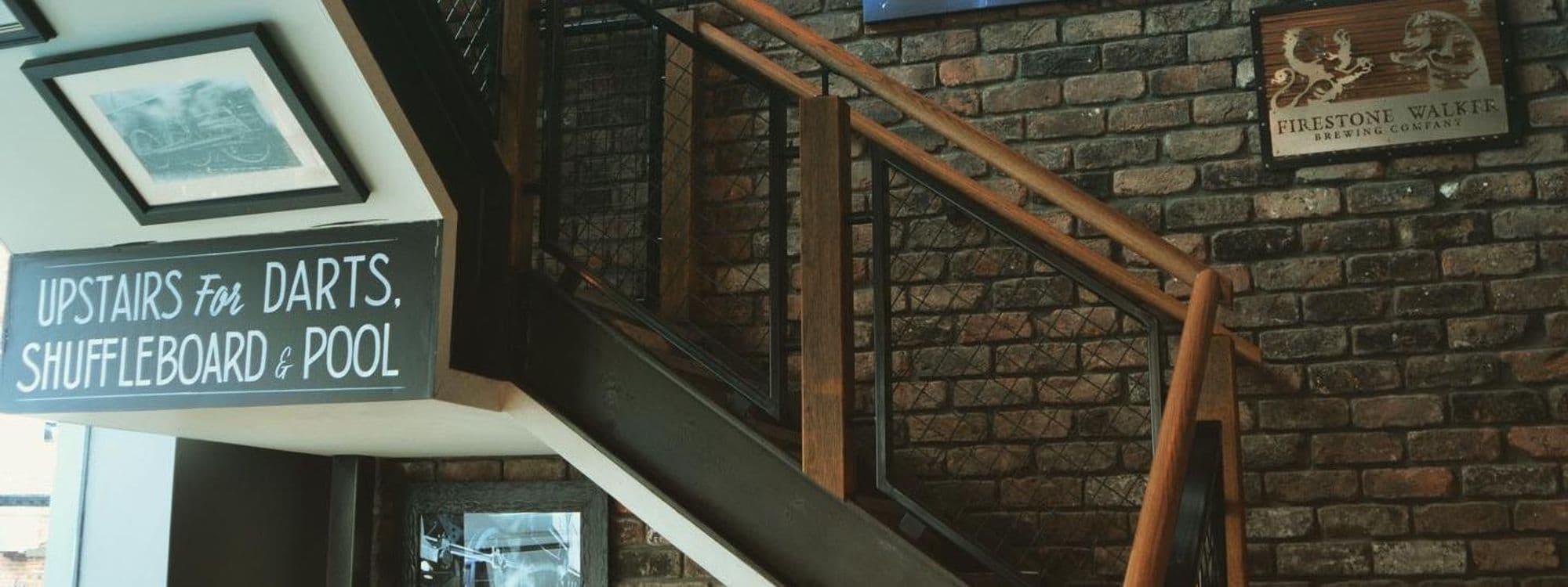 Stairs leading to the upper level at The Head of Steam Headingley, with a sign indicating "Upstairs for Darts, Shuffleboard & Pool." The walls feature exposed brick and framed pictures, creating a cozy atmosphere.
