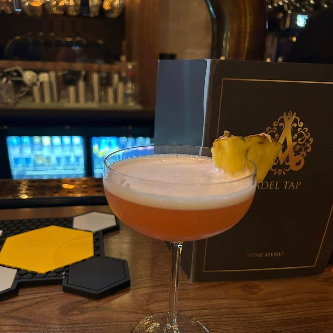 A close-up of a cocktail served in a stylish glass, topped with a slice of pineapple. In the background, there is a wine menu with the title "Adel Tap." The setting features a wooden bar and a colorful display of drinks.