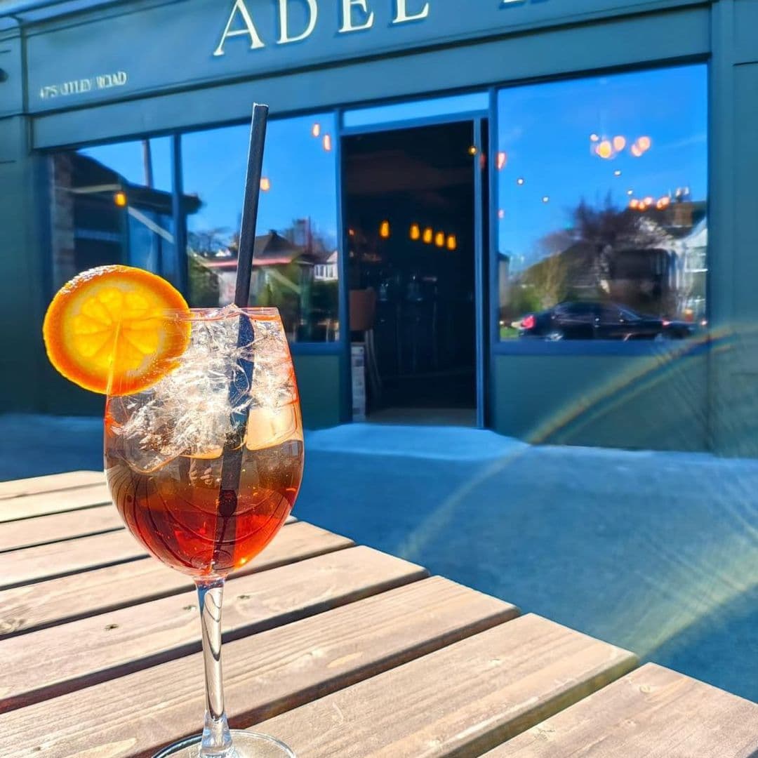 A refreshing drink with ice and a slice of orange sits on a wooden table in front of the entrance to Adel Tap. The building features large windows reflecting a sunny day, with the name "Adel Tap" prominently displayed above the door.