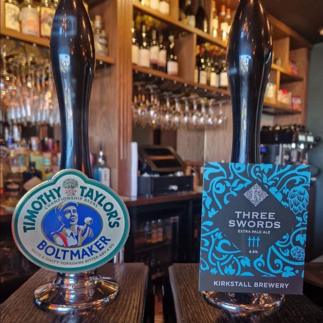 Two beer taps are displayed at a bar in Adel Tap. The left tap features a label for "Timothy Taylor's Boltmaker," a Yorkshire bitter, while the right tap showcases "Three Swords," an extra pale ale from Kirkstall Brewery. The background includes shelves with various bottles and glasses, creating a warm and inviting atmosphere.
