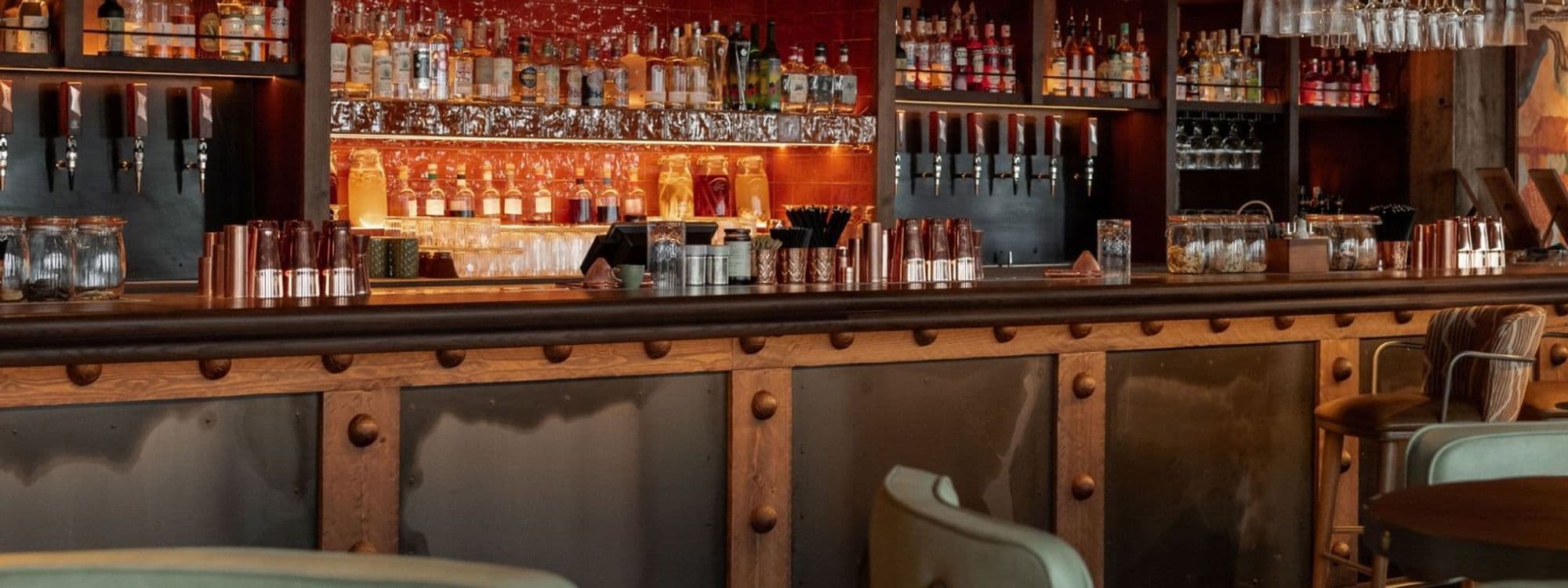 Alt text: A stylish bar at Azotea featuring a wooden counter, shelves lined with various bottles, and glassware. The warm lighting creates a cozy atmosphere, perfect for enjoying drinks.