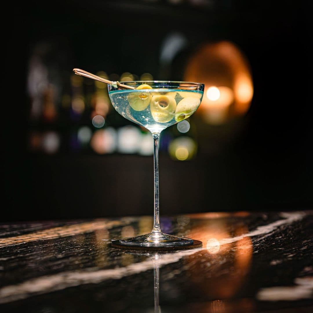 A close-up of a stylish cocktail glass filled with a clear drink and garnished with olives, set on a polished marble surface. The background features softly blurred bar lights, creating an inviting atmosphere. This image represents Dakota Grill.