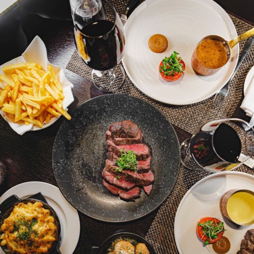 A beautifully arranged table at Dakota Grill featuring a plate of sliced steak garnished with herbs, a side of golden French fries, and various elegant dishes including a tomato with sauce and a creamy mac and cheese. Glasses of red wine complete the dining experience.