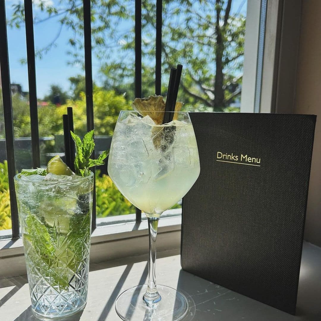 Two refreshing cocktails sit on a windowsill at Deer Park Hotel, with a drinks menu beside them. One cocktail is in a tall glass filled with mint and lime, while the other is in a large round glass with ice and a garnish. Sunlight streams through the window, highlighting the vibrant greenery outside.