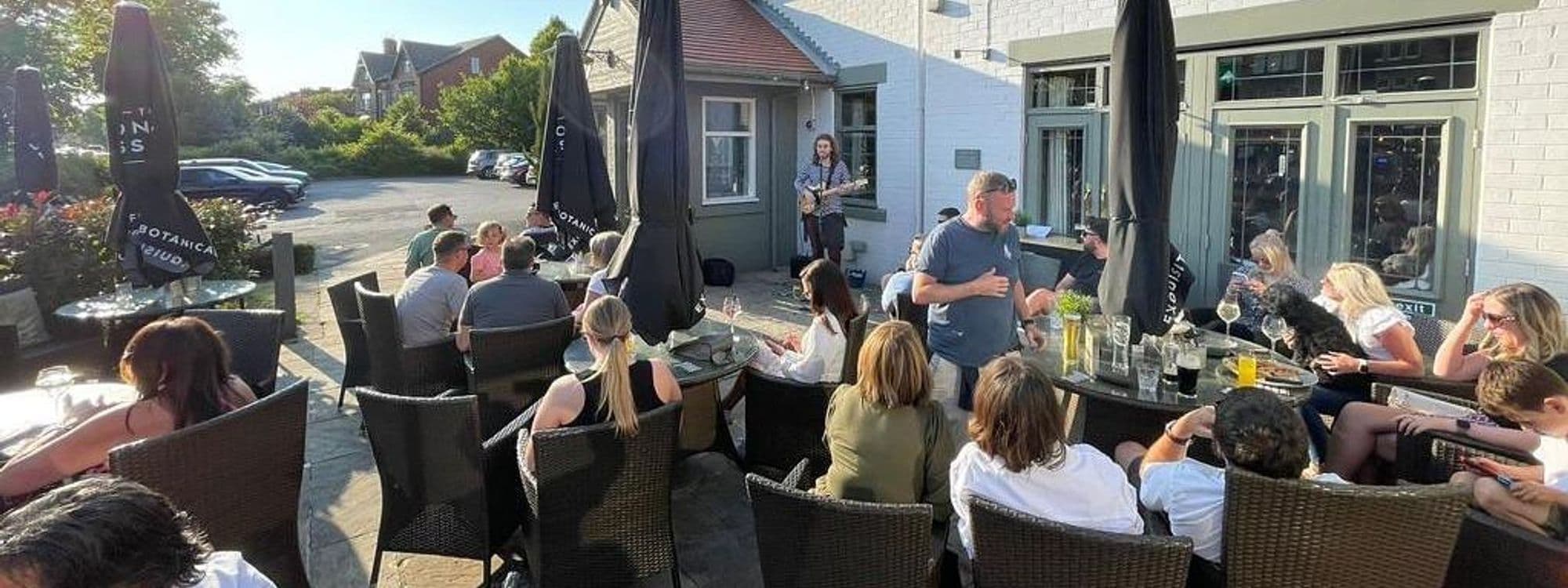 Outdoor seating area at Deer Park Hotel with guests enjoying drinks and live music. People are sitting at tables under large umbrellas, creating a relaxed atmosphere. The scene includes a mix of adults and children, with some pets present, surrounded by greenery.