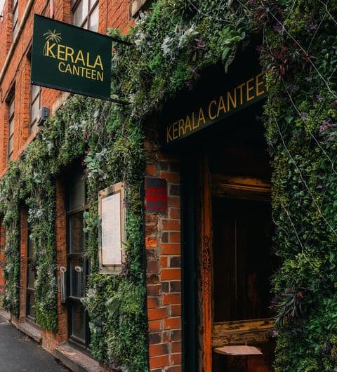 Alt text: The entrance of Kerala Canteen, featuring a brick wall adorned with lush greenery and plants. A sign above the door displays the name "Kerala Canteen" in bold letters.