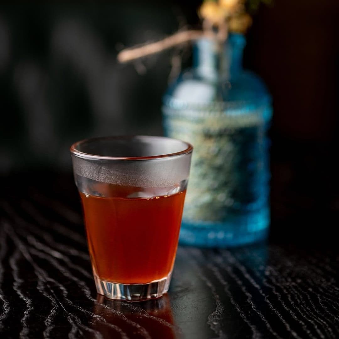 A close-up of a glass filled with dark, aromatic tea, placed on a wooden surface. In the background, a blue glass bottle adds a pop of color, enhancing the cozy atmosphere. This image represents the warm and inviting experience at Kerala canteen.