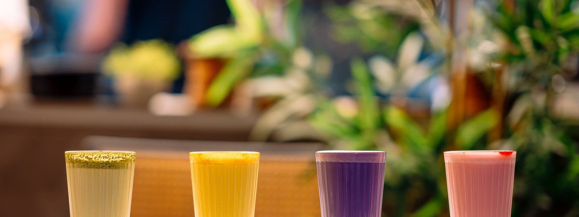 Four colorful drinks in tall glasses are arranged in a row on a table at Nesso Coffee. The drinks include a green, yellow, purple, and pink beverage, with a blurred background of plants and a cozy cafe atmosphere.