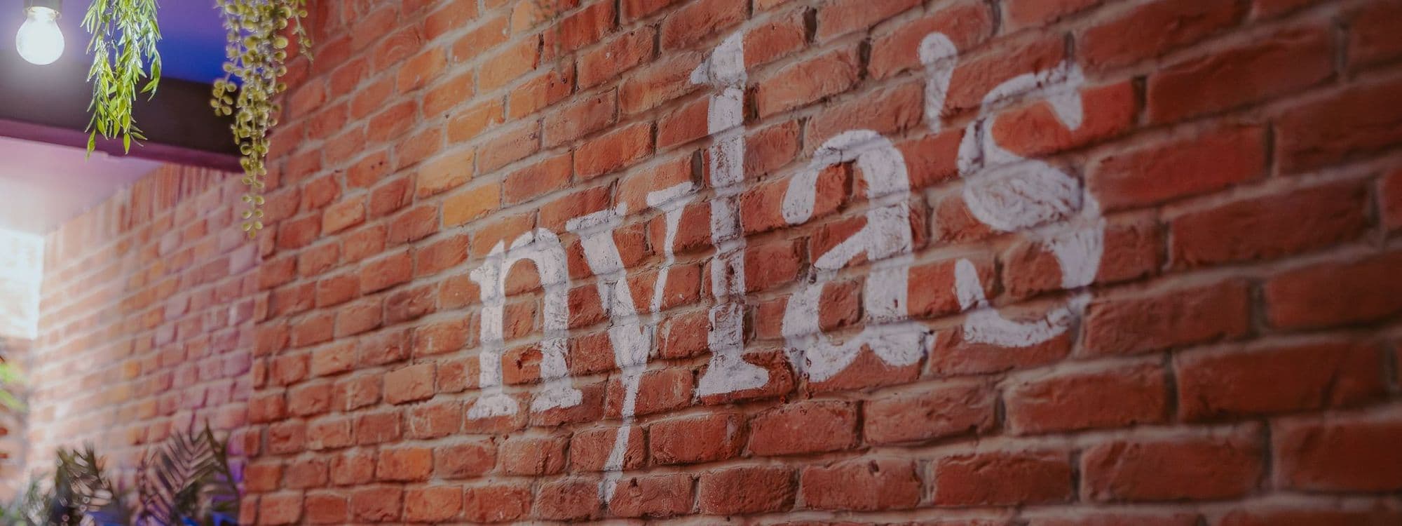 Brick wall with the name "Nyla's" painted in white. Green plants hang from the ceiling, creating a cozy atmosphere. Soft lighting adds warmth to the space.
