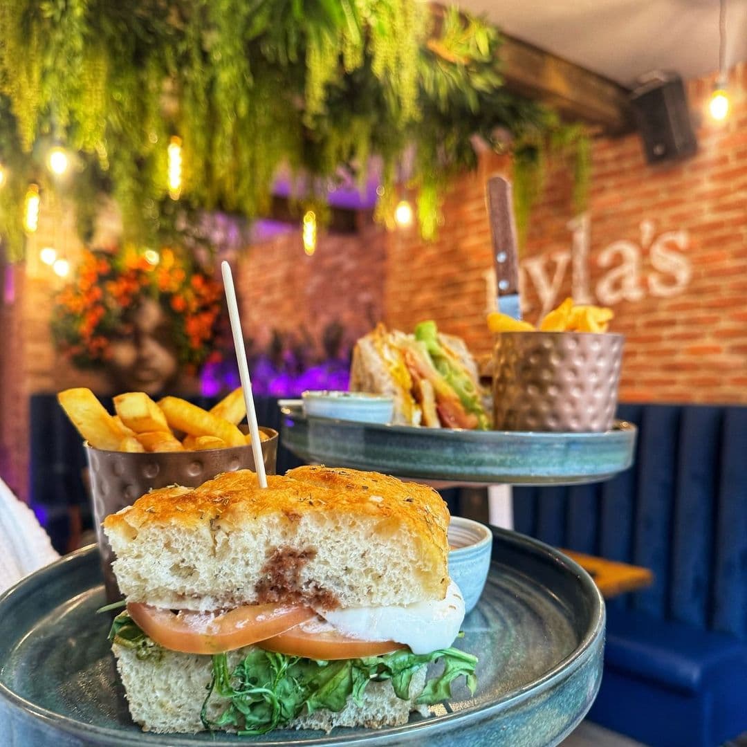 A delicious sandwich with fresh ingredients, including lettuce and tomato, served on a plate at Nyla's. In the background, a portion of golden fries in a cup and another sandwich can be seen on a tiered stand. The restaurant features a cozy atmosphere with greenery and warm lighting.