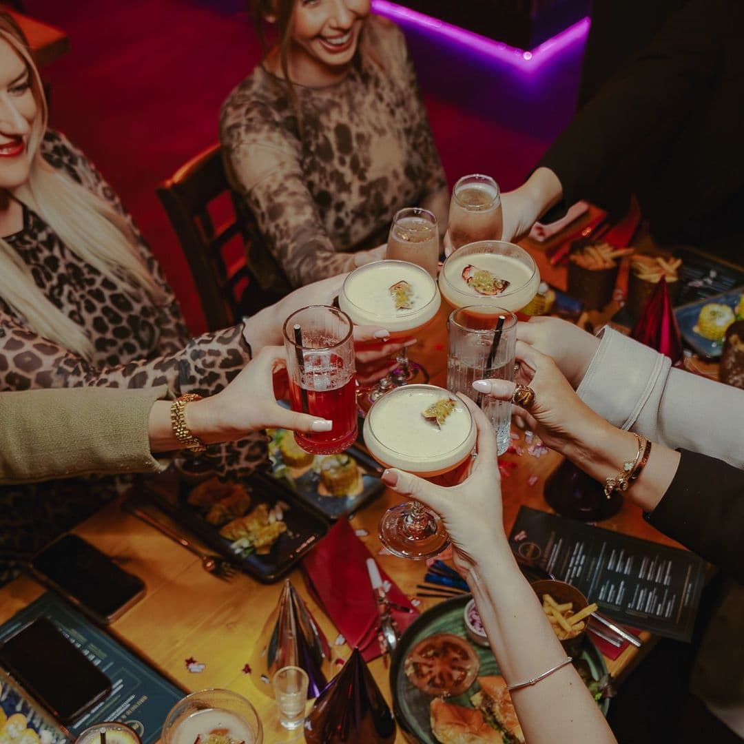 Group of friends toasting with colorful cocktails at Nyla’s. The table is filled with delicious food and festive decorations, creating a lively atmosphere.