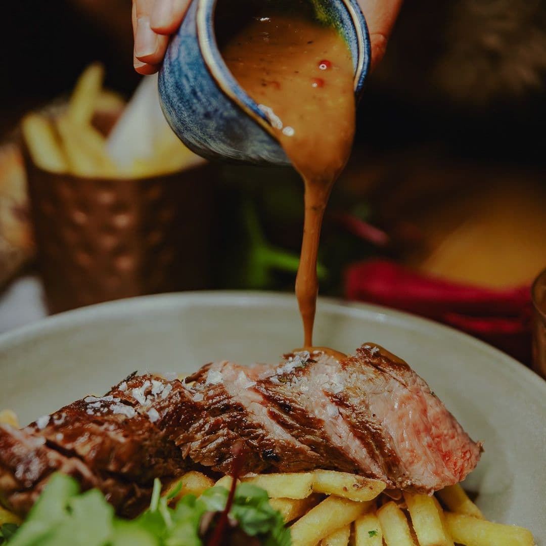 A close-up of a delicious meal at Nyla’s, featuring a juicy piece of steak topped with savory sauce, served alongside golden French fries and fresh greens. A hand is pouring the sauce over the steak, enhancing the dish's appeal.
