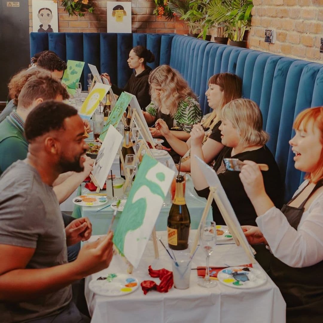 A lively group of people enjoying a painting class at Nyla’s. They are seated at a long table with easels and canvases in front of them. Each person is engaged in painting, with colorful palettes and drinks on the table. The atmosphere is cheerful and creative, with greenery and artwork in the background.