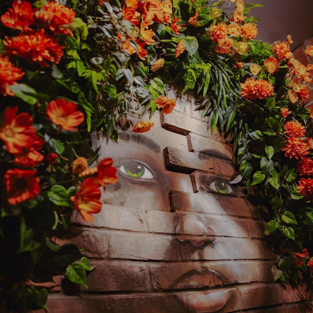 A close-up of a mural featuring a face with striking green eyes, surrounded by vibrant orange flowers and lush green foliage. The artwork is set against a textured brick wall, creating a lively and inviting atmosphere at Nyla’s.