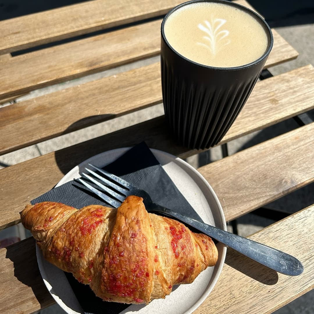 A warm, flaky croissant with a golden crust sits on a plate beside a latte in a textured black cup, on a wooden table.