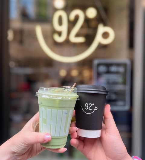 Two hands hold drinks in front of a cheerful 92 Degrees Coffee shop sign, showcasing a green beverage and a coffee cup.