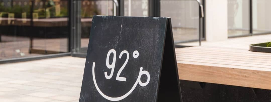 Chalkboard sign with "92°" and a smiling face, set outside a modern coffee shop with large glass windows.