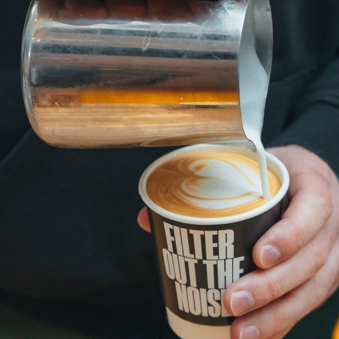 A barista pours steamed milk into a cup, creating a latte art swirl, with a cozy café atmosphere in the background.