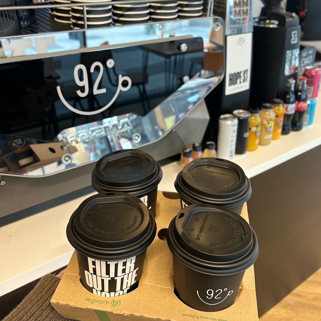 Four black coffee cups in a cardboard carrier sit in front of a sleek coffee machine, with colorful drinks lined up behind.