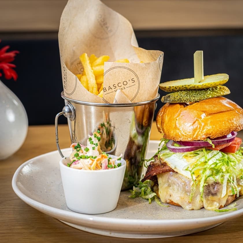Juicy burger with toppings, served with crispy fries in a bucket and a side of coleslaw, set on a rustic table.