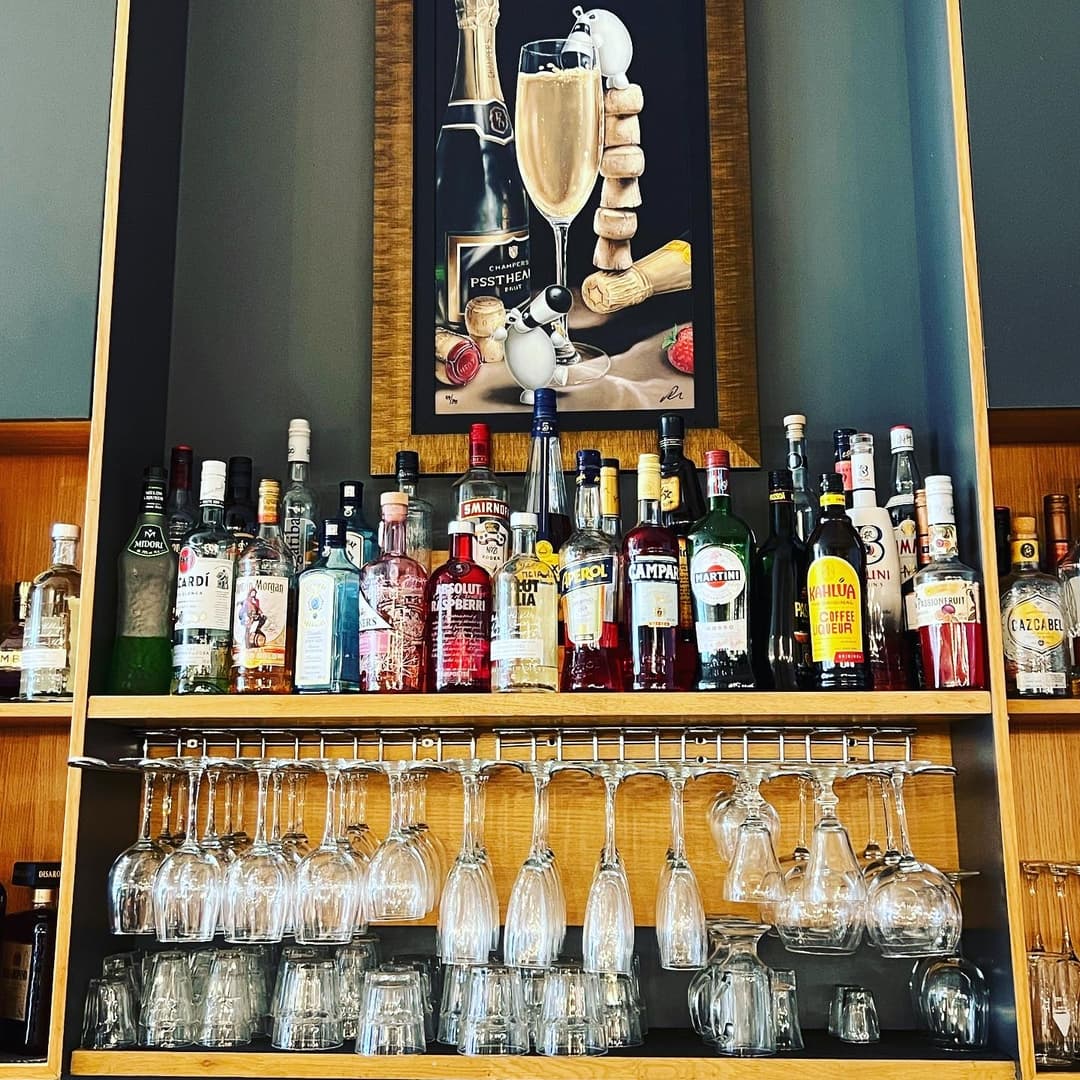 Colorful liquor bottles line a wooden bar shelf, with elegant glassware hanging below and a champagne-themed artwork above.