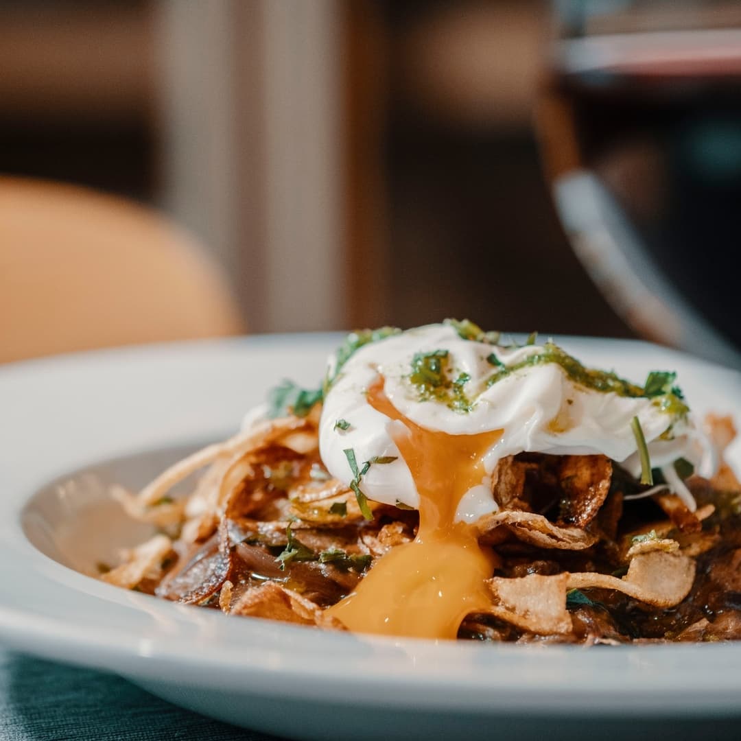 A close-up of a plate featuring crispy potatoes topped with a poached egg, drizzled with herbs and sauce, with a glass of wine in the background.
