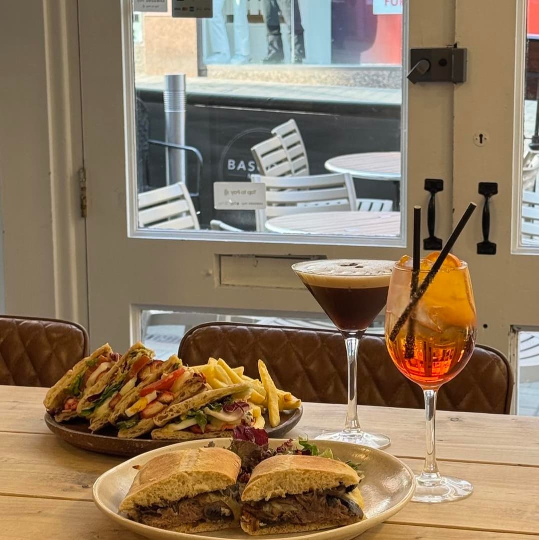 A wooden table displays a hearty sandwich, fries, a cocktail, and a coffee, with outdoor seating visible through the window.